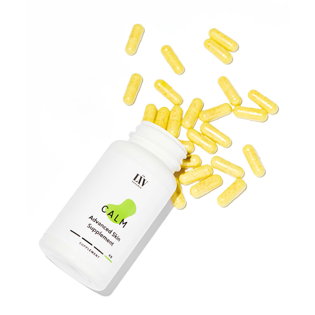 A bottle and box of board certified dermatologist Dr. Liv Kraemers Calm Advanced Skin Supplement, lying on a white surface, letting the yellow capsules roll out.
